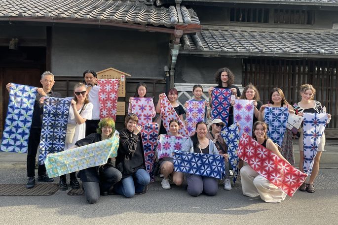 A group of students hold up traditional Japanese cloth in front of a Japanese building